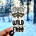 Sticker with butterfly design and 'Wild and Free' text held in front of a snowy forest.