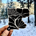 Decorative sticker with a black floral design and white text held in front of a snowy landscape.