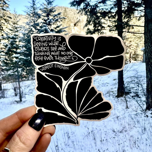 Decorative sticker with a black floral design and white text held in front of a snowy landscape.