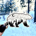 Sticker of a bear with trees inside its body held in front of a snowy forest background