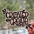Sticker with 'CREATED TO CREATE' text held against a blurred natural background