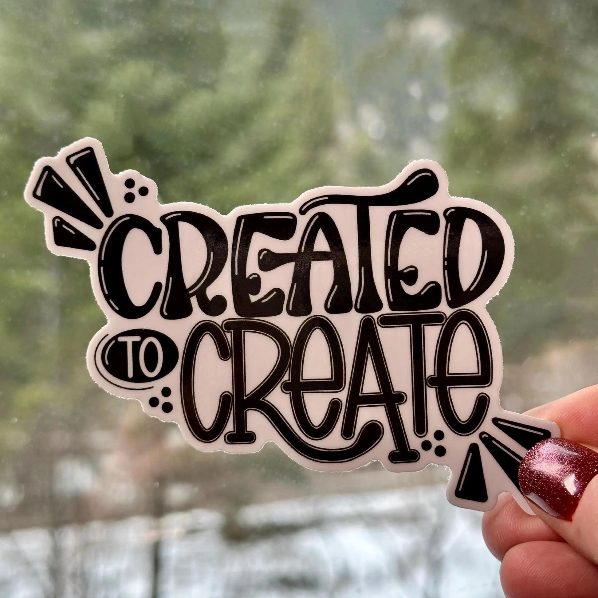 Sticker with 'CREATED TO CREATE' text held against a blurred natural background