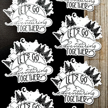 Black and white 'Let's Go Adventuring Together' stickers on a wooden surface.