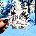 Hand holding a vinyl sticker with 'Let's Go Adventuring Together' text against a snowy forest background