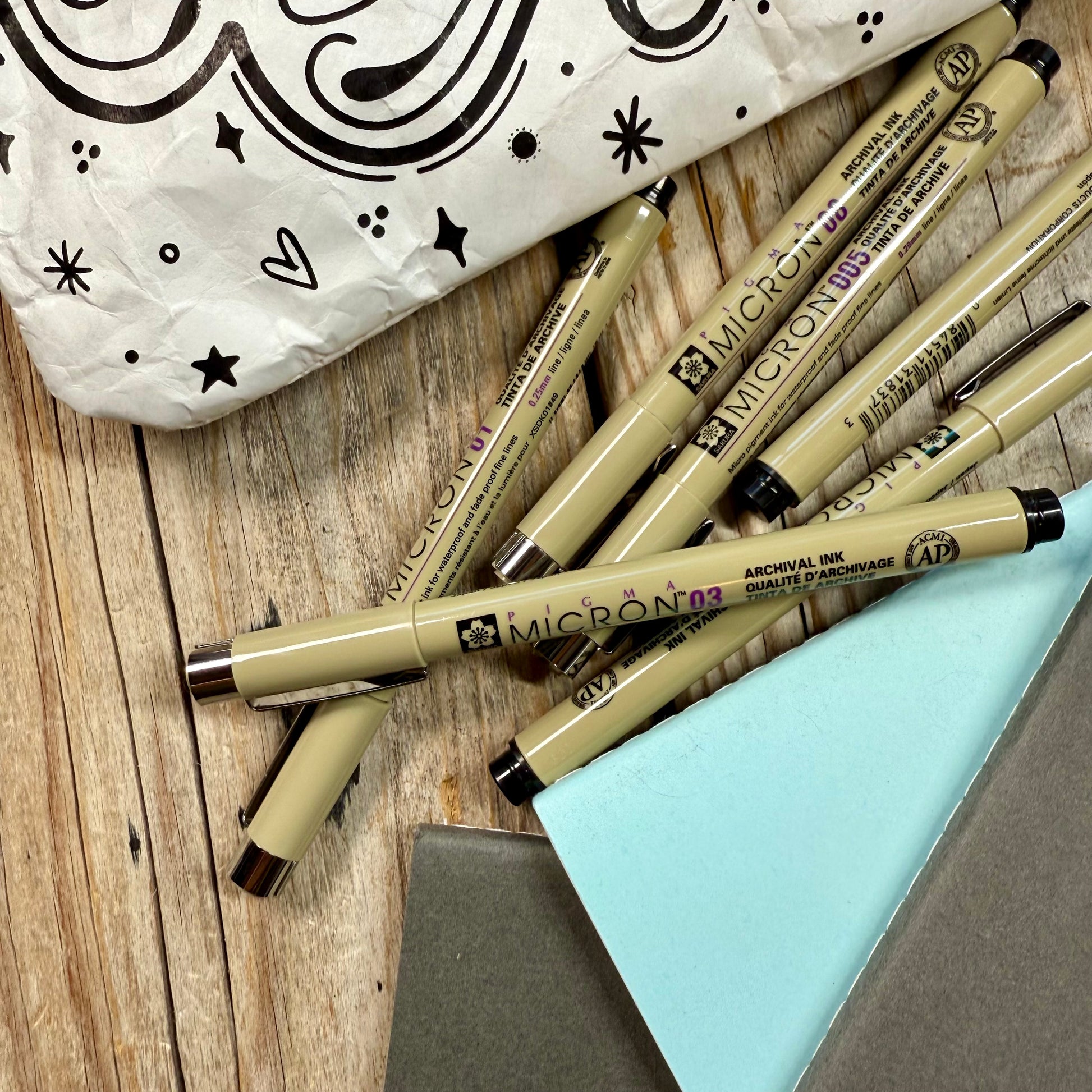 Micron pens arranged beside dotted notebook and lettering workbook on desk