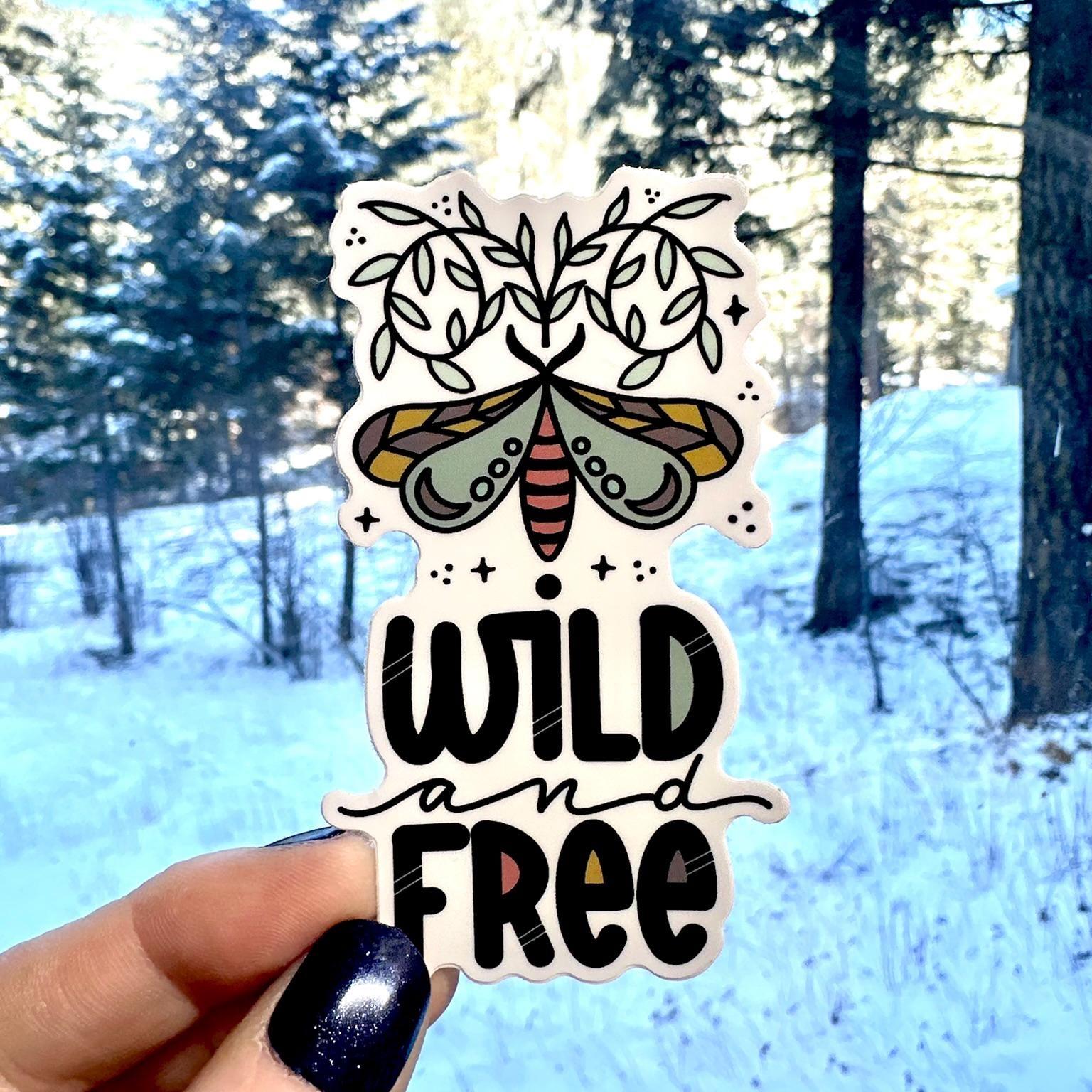 Sticker with butterfly design and 'Wild and Free' text held in front of a snowy forest.