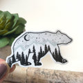 Sticker of a bear with a forest design held by a hand on a wooden surface.