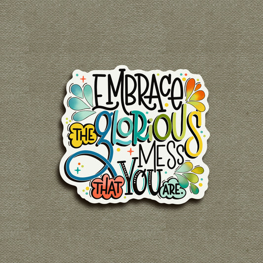 A colorful hand lettered vinyl sticker with the quote 'Embrace The Glorious Mess You Are' on a grey background.